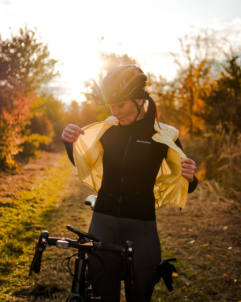 Cyclist pulling on a lightweight yellow SOOMOM Base Wind Gilet on a sunlit gravel path